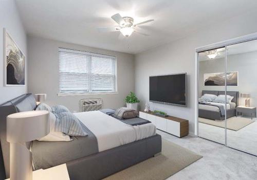 Light grey walls & carpet in bedroom. Mirror closet doors, mounted TV, & window. With A/C unit & fan